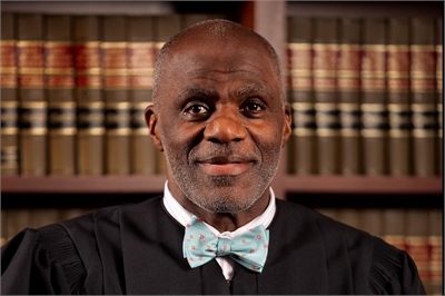 [ai] A smiling judge wearing a black robe and a light blue bow tie, standing in front of a large bookshelf filled with legal books.